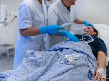 Yatakta oksijen maskesi takmış bir hastanın olduğu hastane sahnesi. Acil servis sırasında yatağın üzerinde su toplayan hemşireler de vardı.