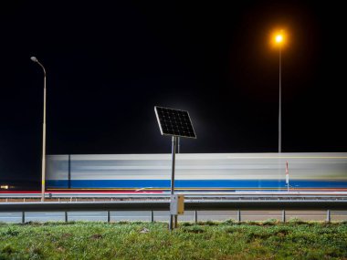 Gece Hollanda otoyolunun kenarına monte edilmiş güneş paneli. Uzun açık kamyonet izleri ve sokak ışıkları renkli çizgiler oluşturuyor.