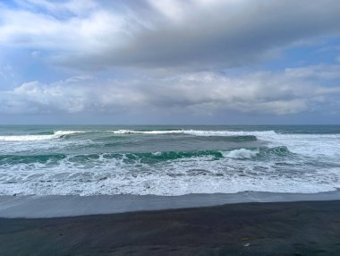 Dramatic dark sand beach features turquoise waves rolling under a cloudy sky, creating a peaceful and natural seascape perfect for travel and environmental marketing.