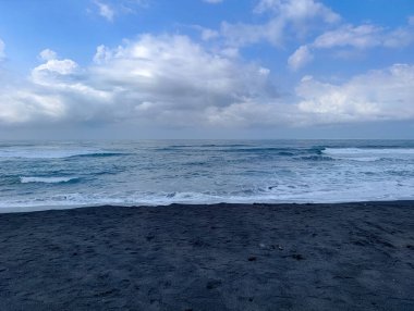 Dark beach meets the wavy turquoise ocean under a cloudy blue sky, creating a serene and peaceful coastal landscape perfect for travel and vacation-related marketing.