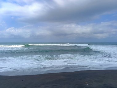 Ocean waves are crashing onto the black sand beach under a cloudy blue sky, creating a peaceful and dramatic seascape ideal for travel or nature-themed visuals.