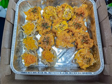 Savory dim sum sit appetizingly in a foil container, some topped with bright orange sauce and shredded vegetables, perfect for food-related promotions and Asian cuisine designs.