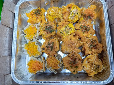 Freshly prepared dumplings with orange and yellow sauces, sprinkled with herbs, sit in a foil tray in a top-down composition, ideal for food service or catering advertising.