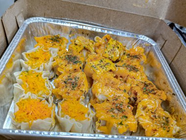 Freshly made dumplings, some topped with shredded orange filling and others with a creamy orange sauce and herbs, presented in a foil tray inside a cardboard box, creating an appetizing visual perfect for food-related marketing.