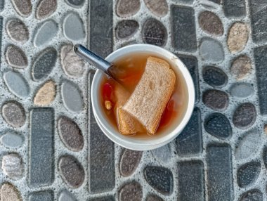 Sweet brown soup with bread pieces rests in a white bowl beside a spoon on a grey stone surface, creating a simple and casual composition suitable for food blogs and cultural content.