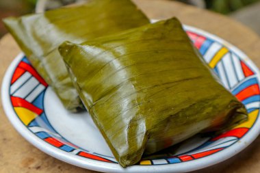 Yeşil yapraklara sarılı iki yiyecek renkli bir tabakta oturmakta. Keyfini çıkarmaya hazır. Yemek, kültür ve mutfak temalarını öneriyor..