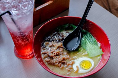 Çorba, et, yumurta ve yeşilliklerle dolu kırmızı bir ramen kasesinin genel manzarası, büyük bir bardak kırmızı köpüklü içeceğin yanında, restoran ve yemek promosyonları için uygun bir aşçılık sahnesi yaratıyor..