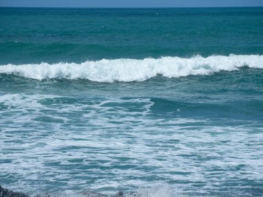 Köpüklü deniz mavisi dalgaları, parlak gün ışığında beyaz köpük ve dokularla kırılıyor. Seyahat ve çevre pazarlaması için uygun sakin bir kıyı manzarası yaratıyor..