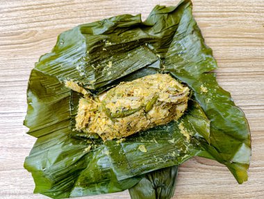 Bitkilerle buharlanmış balık tabağı açık kahverengi ahşap bir masanın üzerinde yeşil muz yapraklarına sarılı olarak yemek blogları ve mutfak promosyonları için ideal iştah açıcı, organik bir sunum oluşturur..