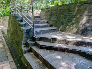 Yosun kaplı tuğla duvar boyunca yükselen metal korkuluklu taş merdiven yemyeşil parkta doğa ve manzara için mükemmel bir açık hava manzarası yaratıyor..