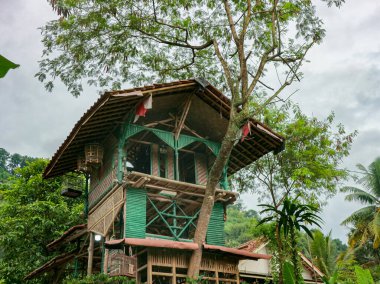 Yeşil aksanlı ve bambu korkuluklu bir kır evi, bulutlu gökyüzü ile tropikal bir orman ortamında yemyeşil ağaçların arasına inşa edilir. Çevre turizmi ve açık havada yaşamak için ideal barışçıl ve maceraperest bir atmosfer yaratır..