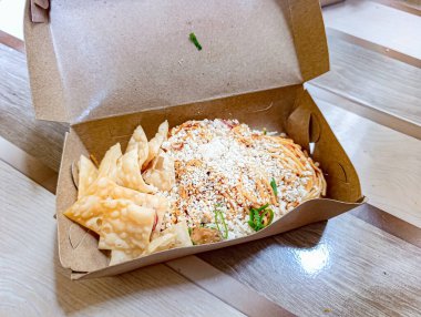 Ahşap masa üzerinde kahverengi bir karton kutuda gevrek wontonlar bulunan Asya eriştesi tabağı. İçinde süslemeler ve serpiştirilmiş peynir var. Günlük ve iştah açıcı bir atmosfer yaratıyor. Yemek dağıtımı için mükemmel bir ortam..