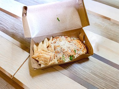 İçinde rendelenmiş peynirli spagetti ve ahşap bir masada kızarmış wonton bulunan bir paket. Doğal ışıkta yakalanmış. Yemek teslimatı ya da restoran pazarlaması için mükemmel sıradan ve iştah açıcı bir ortam yaratıyor..