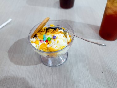 Açık renkli bir masada şeffaf cam bir tabakta rengarenk, kurabiye ve mısır gevreği parçalarıyla kaplı kremalı dondurma yemek ve tatlı pazarlama için mükemmel, neşeli ve davetkar bir tatlı sahnesi yaratır..