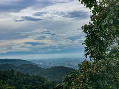 Yoğun ağaçlarla dolu yemyeşil bir dağ manzarası ve bulutlu bir gökyüzünün altında yuvarlanan tepeler seyahat ve açık hava macerası için ideal barışçıl ve doğal bir atmosfer yaratıyor..