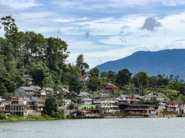 Bereketli bir yamaçta evleri olan sakin bir su kütlesinin yanında yeşil ağaçlar ve dağlarla çevrili parçalı bulutlu bir gökyüzü altında seyahat ve doğa pazarlaması için ideal bir köy..