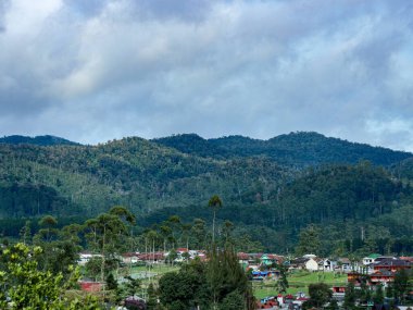 Yoğun yeşil ormanlar ve bulutlu gökyüzü altında evleri olan küçük bir köyün bulunduğu manzaralı dağ manzarası seyahat ve doğa temaları için mükemmel barışçıl ve doğal bir atmosfer yaratıyor..