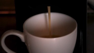 Kahve makinesinden beyaz bir seramik fincana kahve dökülüyor. Karanlık arka plan demleme sürecini ön plana çıkarıyor. Sabah rutinleri, içecekler ve kahve kültürü için editoryal kullanım için ideal.