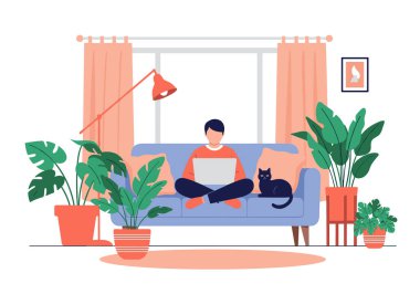 Genç bir adam evdeki rahat bir kanepede otururken dizüstü bilgisayarda çalışıyor ve bir sürü ev bitkisinin arasında siyah kedisi var..