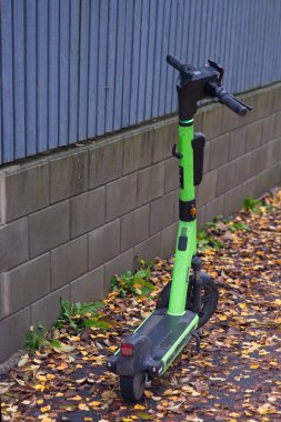Yeşil elektrikli scooter, devrilmiş sonbahar yapraklarıyla çevrili yuvarlak metal duvarın yanında kaldırıma park edilmiş.,