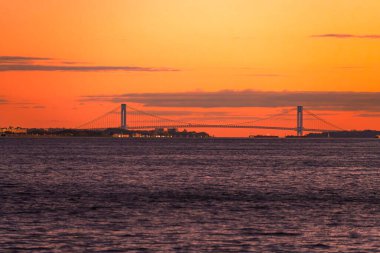 Verrazzano-Narrows Köprüsü, aşağıdaki sularda gemilerin geçtiği canlı bir günbatımı gökyüzüne karşı aydınlanıyor..