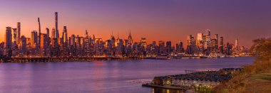 New York şehrinin ufuk çizgisi gün batımında parlıyor, Hudson Nehri alacakaranlık gökyüzünün canlı renklerini yansıtıyor..