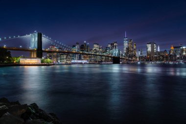 İkonik Brooklyn Köprüsü gece gökyüzüne karşı aydınlanıyor. Arka planda Manhattan silueti parıldıyor..