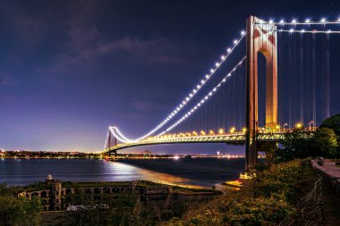 Verrazano 'nun gece parlaması New York' taki Brooklyn ve Staten Island 'ı birbirine bağlayan Verrazano-Narrows Köprüsü' nü aydınlatıyor. Fort Wadsworth, 19. yüzyılda inşa edilmiş, ön planda duruyor..