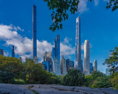 New York şehrinin ikonik silueti Central Park 'ın üzerinde yükseliyor, açık mavi gökyüzü altında modern gökdelenler ve yemyeşil bir karışım sergiliyor..