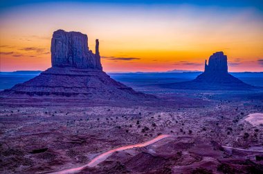 Valley Sunset Road Trip: Görüntü ikonik Anıt Vadisi 'ni alacakaranlıkta çekiyor, Mittens silueti canlı, renkli bir gökyüzüne karşı. Bir arabanın farları çöl zemininde çizgiler çizer, hareket hissi verir ve sonsuzluğu vurgular.