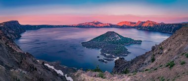 Oregon 'daki Krater Gölü Ulusal Parkı günbatımının pembe tonlarıyla yıkanırken, Büyücü Adası, volkanik kül konisi, kalderadan yükseliyor. Büyücü Adası, kalderayı oluşturan patlamadan sonra oluştu ve parkın eşsiz jeologuna eklendi.