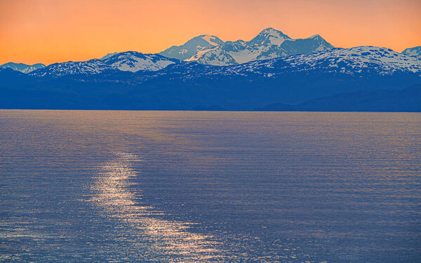 Солнце садится за заснеженные горы, бросая оранжевое свечение над небом и размышляя о спокойной воде.