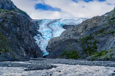 Engebeli Alaska manzarası boyunca uzanan buzulun buzlu mavi yüzeyi karanlık, kayalık dağlarla ve aşağıdaki çalkantılı nehirle çelişiyor..
