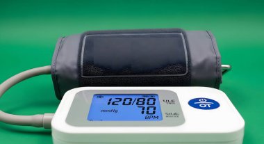 A close-up shot of a digital blood pressure monitor with a cuff and a display showing a reading of 12080, set against a plain green backdrop.