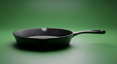 A single, empty cast iron frying pan is presented against a solid green backdrop, highlighting its durable construction and timeless appeal for culinary use.