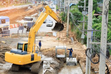 Büyük sarı bir kazıcı bir yol inşaatındaki hendeğe büyük bir beton boru yerleştiriyor. Bu ağır makineler, yeni bir altyapı projesi kapsamında yeraltı drenajı veya su sistemleri kurmak için kullanılır..