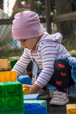 Yeni yürümeye başlayan bir çocuk, açık hava oyunları sırasında büyük renkli bir blok yerleştirirken konsantrasyon içinde çömeldi. Odaklanma, ince motor beceriler ve keşif üzerine vurgu.