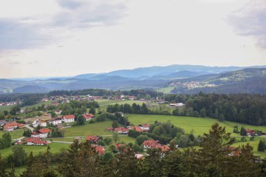 Dağılmış evleri, yeşil çayırları ve uzak mavi dağları olan huzurlu kırsal alan. Bulutlu bir gökyüzünün altında sakin ve uyumu simgeleyen pitoresk bir kır manzarası..