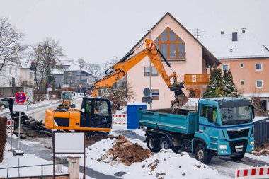 Sarı kazıcı, karlı bir caddede bir çöp kamyonuna toprak taşıyor. Arka planda konut ve inşaat bariyerleri görünüyor..