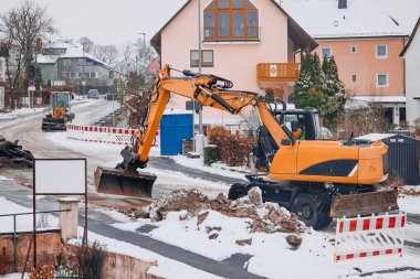 Kazıcı, kış inşaat çalışmaları sırasında karlı bir yerleşim bölgesindeki toprağı kazıp kaldırıyor. Bölgenin etrafında kar yığınları ve güvenlik engelleri.