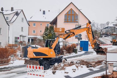 Karlı bir kentsel yol çalışması sahasında geçici yol bariyerleri ve tabelaları yakınında çalışan kazıcının geniş görünümü, alan düzeni ve güvenlik tedbirlerini gösteriyor.