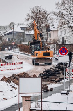 Yol tadilatı sırasında karlı bir sokakta çalışan bir kazıcı. Asfalt atıkları, güvenlik bariyerleri ve kış koşulları aktif inşaat mühendisliği çalışmalarını gösteriyor.