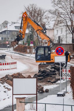 Karlı bir yerleşim bölgesinde yol yenileme sırasında ağır kazıcı asfaltı kepçeledi. İnşaat engelleri ve kış koşulları altyapı çalışmalarının devam etmekte olduğunu vurguluyor.