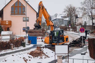 Sarı bir kazıcı, yerleşim alanındaki yol onarımı sırasında asfalt moloz yığınlarını temizler. Kar, inşaat bariyerleri ve yıkılan kaldırımlar aktif altyapı bakımını vurguluyor.