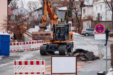 Güçlü bir kazıcı, kış yıkımı sırasında asfalt parçaları kırıyor ve hareket ettiriyor. Etrafı güvenlik bariyerleri ve inşaat atıklarıyla çevrili kentsel bir sokakta çalışıyor..