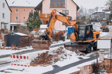 Bir kazıcı, arka planda bariyerler ve konut evleri bulunan kış sokağı tamiri sırasında karlı bir inşaat sahasına toprak taşıyıp temizler..
