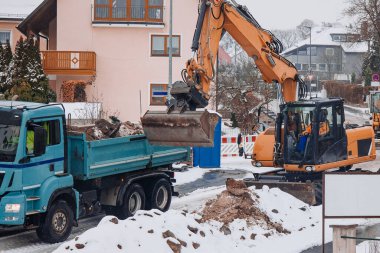 Bir kazıcı kış yollarında kar, güvenlik bariyerleri ve inşaat ekipmanlarıyla çevrili bir kamyona toprak ve enkaz yükler..