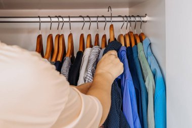 Düzenli bir gardırobun içindeki çeşitli mavi ve gri tonlarda düzenlenmiş gömlekleri karıştıran bir el..