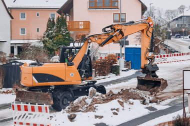 Karla kaplı bir yerleşim bölgesinde sokak yol çalışması yapan inşaat kazıcısı. Kentsel bakım, altyapı onarımı ve kışın ağır ekipmanlar.