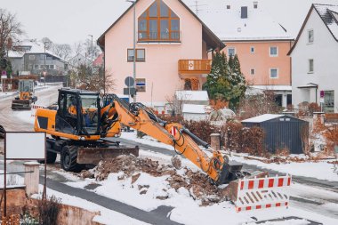Turuncu kazıcı kış boyunca yerleşim yerlerinde bir yol inşaatında çalışıyor. Kentsel altyapı onarımı, sokak bakımı, yol çalışmaları ve ağır makine konsepti.